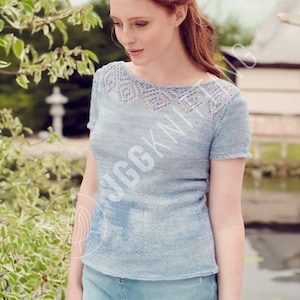 Può includere: Un top in maglia azzurra con maniche corte e scollatura traforata decorativa. Il top ha uno scollo tondo e un sottile motivo a righe. La modella indossa il top all'aperto vicino a uno specchio d'acqua.