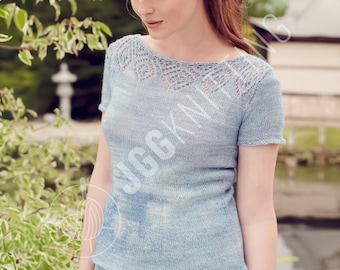 May include: A light blue knit top with short sleeves and a decorative openwork yoke. The top is worn by a person with red hair, standing outdoors near greenery and water. The top is a soft, light blue color.