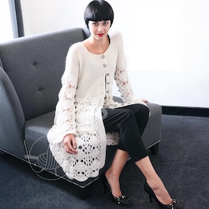 Könnte beinhalten: Ein cremefarbener Cardigan mit offener Front und aufwendigen Spitzendetails. Der langärmlige Pullover hat dekorative Knöpfe und einen gewellten Saum. Das Model trägt eine schwarze Hose und Absätze und sitzt auf einem grauen Sessel.