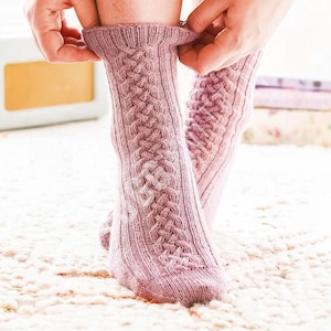 Peut inclure: Paire de chaussettes en tricot de câble rose clair. Les chaussettes présentent un motif de câble détaillé sur le devant et des poignets côtelés. Les chaussettes sont ajustées par les mains d'une personne. Sur une surface blanche texturée.