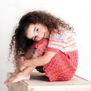 May include: A young person wearing a knitted coral-colored dress and top with a striped pattern. The dress has a textured design. The person is sitting on a wooden box, with their arms wrapped around their legs.