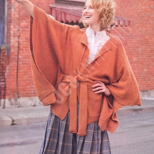 May include: An orange-brown knit wrap with a matching belt. The wrap has wide sleeves and a shawl collar. The person is wearing a white blouse and a gray plaid skirt. The background is a brick wall.
