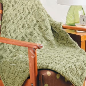 May include: A sage green knitted blanket with a diamond pattern is draped over a wooden chair. The blanket's texture is visible, and it appears soft and cozy. The chair has a brown seat with gold polka dots.