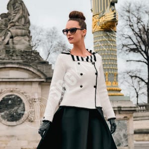 Może przedstawiać: Biała kurtka z czarnymi detalami, w tym w paski kołnierzem i mankietami oraz czarnymi guzikami. Kurtka jest noszona z czarną spódnicą. Zestaw uzupełniają czarne rękawiczki i okulary przeciwsłoneczne, tworząc elegancki, ponadczasowy wygląd.
