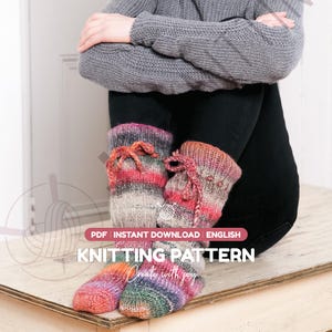 May include: A pair of hand-knitted socks featuring a variegated yarn pattern in shades of pink, orange, grey, and green. The socks have a ribbed cuff with a decorative braided tie. The image includes text: PDF INSTANT DOWNLOAD ENGLISH KNITTING PATTERN Create with joy.