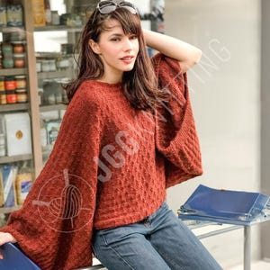 Könnte beinhalten: Ein rostfarbener Strickponcho mit weiten Ärmeln. Der Poncho hat ein strukturiertes Muster und einen runden Ausschnitt. Das Model trägt eine blaue Jeans und eine Sonnenbrille auf dem Kopf. Der Hintergrund zeigt ein Geschäft mit verschiedenen Artikeln.