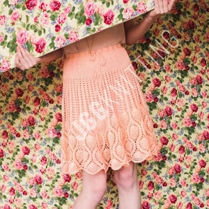 Pode incluir: Uma saia de crochê cor de pêssego com cintura com cordão e bainha com vieiras. A saia apresenta um design vazado com padrões em forma de coração ao longo da borda inferior. O fundo é um padrão floral com rosas cor-de-rosa sobre um fundo amarelo.