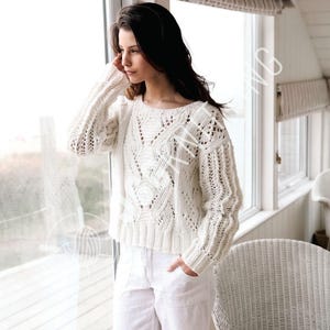 May include: A cream-coloured, open-weave sweater with a round neckline and long sleeves. The sweater features a textured cable knit design on the front and sleeves. The model is wearing white trousers.
