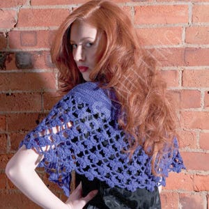May include: A blue crochet shawl with a floral pattern. The shawl is draped over the shoulders. The background is a red brick wall.