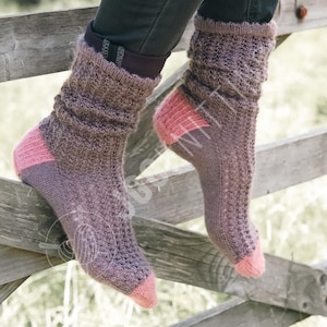 May include: A pair of knitted socks with a textured pattern. The socks are primarily brown with pink accents on the toes and heels. The cuffs are scrunched down. The socks are resting on a wooden fence.