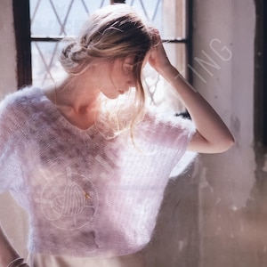 Puede incluir: Un suéter rosa claro y transparente de manga corta con cuello en V. El suéter tiene un aspecto texturizado y difuso y un gráfico blanco en la parte delantera. La imagen está suavemente iluminada, con una persona en el fondo.