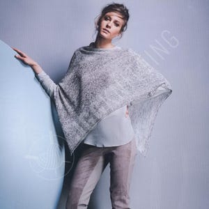 May include: A light gray knitted poncho with a textured pattern. The poncho is draped over a person wearing a light gray long-sleeved shirt and brown pants. The background is a light blue and gray.