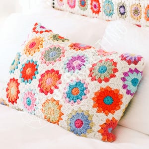 Puede incluir: Un cojín cuadrado de ganchillo con un diseño floral en varios colores, incluyendo naranja, rosa, azul y verde. El cojín está hecho de hilo blanco y presenta un motivo repetido de flores coloridas. Un cojín a juego alargado es visible en el fondo.