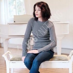 Op de afbeelding: Een grijze gebreide trui met lange mouwen. De trui heeft een geribbelde textuur en wordt gecombineerd met een donkerblauwe jeans. De persoon zit op een bankje, met een witte piano op de achtergrond.