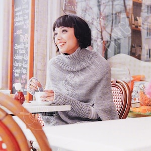 Könnte beinhalten: Eine Frau trägt einen grauen Strickponcho mit hohem Kragen und sitzt an einem Cafétisch. Sie lächelt und hält eine Tasse Kaffee. Der Hintergrund umfasst eine Menütafel und ein Fenster mit einer verschwommenen Außenszene.