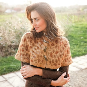 May include: A brown and tan knitted cape with a spotted pattern and two wooden toggle closures. The cape is worn over a brown long-sleeved top. The image is taken outdoors in natural light.