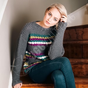 Może przedstawiać: Szary sweter z dzianiny z okrągłym dekoltem i długimi rękawami. Sweter ma poziome paski w kolorze turkusowym, fioletowym, żółtym i białym na piersi. Modelka ma na sobie niebieskie dżinsy i siedzi na drewnianych schodach.