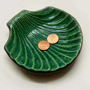 May include: A glossy, deep green ceramic dish shaped like a scallop shell with a ribbed texture. Two copper-colored United States one-cent coins are resting in the center of the dish, providing a sense of scale for the small trinket bowl.
