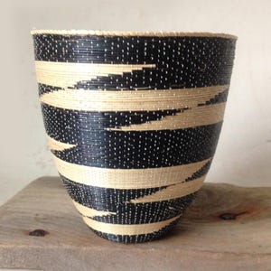 May include: A handcrafted, woven basket with a black and natural color scheme. The basket features horizontal bands of black and beige, creating a striped pattern. The basket is cylindrical with a slightly flared top.