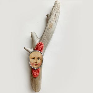 May include: A piece of weathered driftwood adorned with a central oval-shaped portrait of a face. The portrait is framed in silver and flanked by two textured, coral-colored floral embellishments. The driftwood has a light gray, smooth finish.
