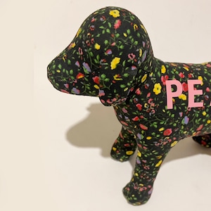 May include: A stuffed dog toy with a black floral pattern featuring pink, yellow, and red flowers. The word "PET" is printed in pink across the dog's back. The toy is on a white surface.