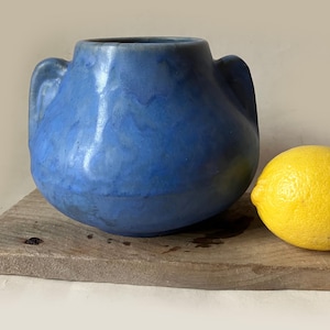 May include: A rounded blue ceramic vessel with two side handles and a mottled glaze finish, positioned next to a single bright yellow lemon. The vessel features a wide opening and a smooth, curved body with subtle variations in the blue color tone.