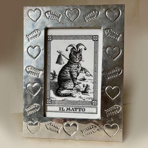 May include: A rectangular silver-toned metal picture frame featuring an embossed border pattern of alternating hearts and fish skeletons. The frame holds a black and white illustration of a cat wearing a jester hat with the text IL MATTO.