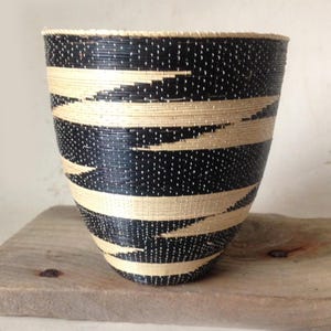 May include: A woven basket featuring a tapered, conical shape with a repeating pattern of horizontal black and cream-colored bands. The exterior surface displays a fine, textured weave throughout, creating a consistent visual rhythm across the entire body.