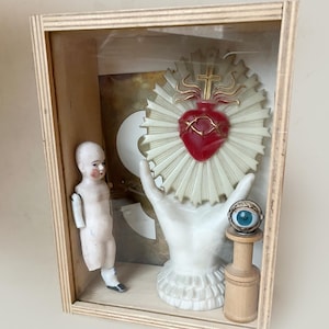 May include: A wooden shadow box containing a porcelain doll, a white hand holding a Sacred Heart, and a wooden stand with a blue eye. The Sacred Heart is red with gold accents. The box has a clear front.