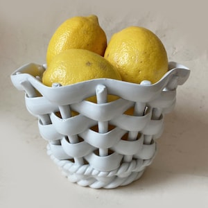 May include: A white ceramic basket with a woven lattice design and a twisted rope-style base, filled with three bright yellow lemons. The bowl features a scalloped, wavy rim and a glossy finish, showcasing a structured, open-weave construction.