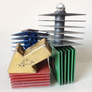 May include: A collection of vintage electronic components featuring stacked metal cooling fins in red, green, blue, and grey. A gold-painted metal arrow-shaped piece rests on top of the red stack, while a bolt secures the top of the grey stack.