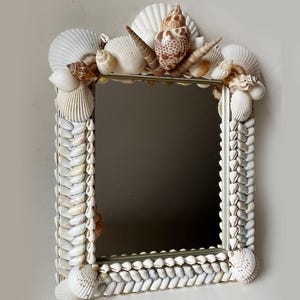 May include: A rectangular mirror with a frame decorated with various white and tan seashells. The top edge features larger, prominent shells, while the sides and bottom are lined with rows of small, uniform white shells arranged in a pattern.