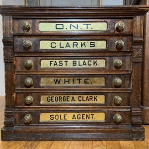 May include: Antique wooden cabinet with six drawers, each labeled with gold lettering. The labels read "O.N.T.", "CLARK'S", "FAST BLACK.", "WHITE,", "GEORGE A. CLARK", and "SOLE AGENT.". The cabinet has brass knobs and is in a well-worn condition.