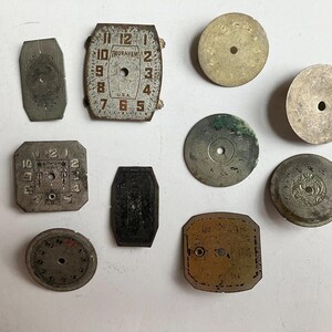 May include: A collection of ten vintage metal watch dials and components in various shapes, including circular, rectangular, and octagonal forms. Several pieces feature printed numerals, decorative engravings, and the text Ingraham U.S.A.