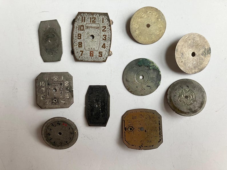 May include: A collection of ten vintage metal watch dials and components in various shapes, including circular, rectangular, and octagonal forms. Several pieces feature printed numerals, decorative engravings, and the text Ingraham U.S.A.