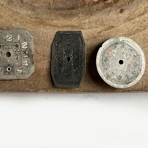 May include: Three vintage watch dials in varying shapes including a square, an elongated hexagon, and a circle. Each metallic dial features engraved numerals and central holes for hands, with the square dial displaying the text CADET and SWISS.