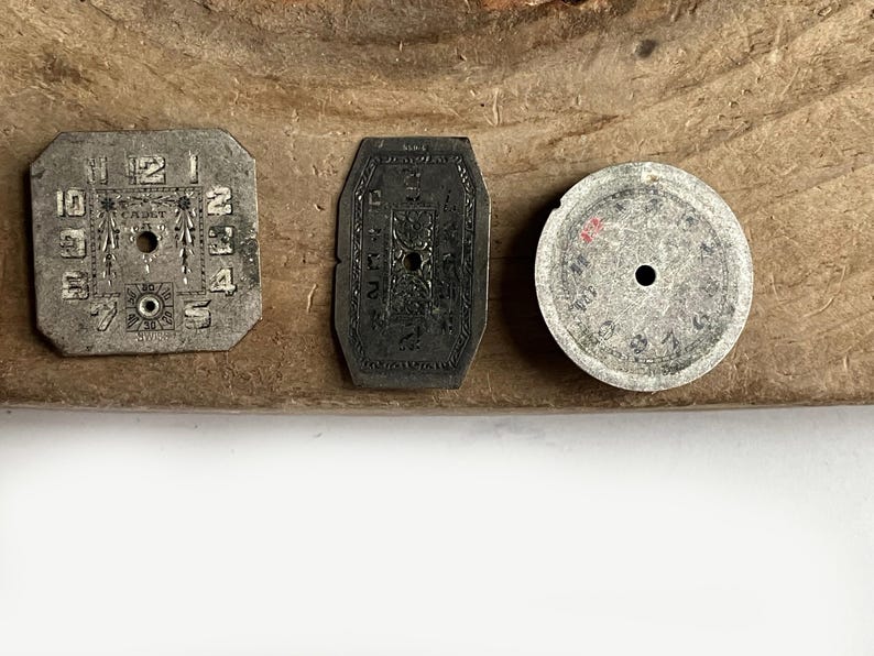 May include: Three vintage watch dials in varying shapes including a square, an elongated hexagon, and a circle. Each metallic dial features engraved numerals and central holes for hands, with the square dial displaying the text CADET and SWISS.