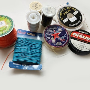 May include: A collection of jewelry making supplies including a spool of bright orange cord, a card of turquoise waxed cotton cord, and various spools of thread and elastic cord labeled Stretch Cord, Magic, and FireLine Crystal braided bead thread.