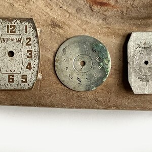 May include: Three vintage watch components including a rectangular Ingraham watch face with numerals 1 through 12 and U.S.A. text, a circular metallic disc with green oxidation, and a small rectangular silver-toned metal plate with floral engraving.