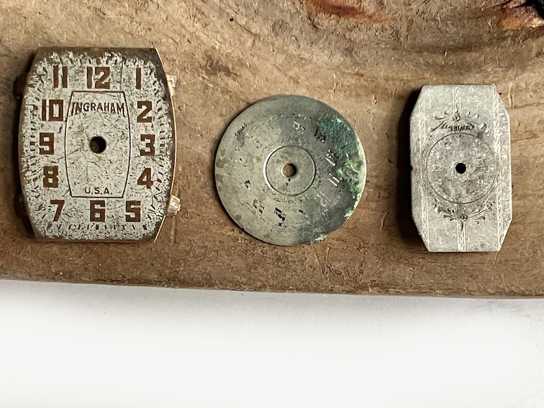 May include: Three vintage watch components including a rectangular Ingraham watch face with numerals 1 through 12 and U.S.A. text, a circular metallic disc with green oxidation, and a small rectangular silver-toned metal plate with floral engraving.