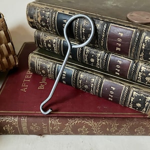 May include: A silver-colored metal boot hook with a circular handle and a curved tip, positioned against a stack of three antique leather-bound books with gold-embossed lettering and a single copper coin resting on the top cover.