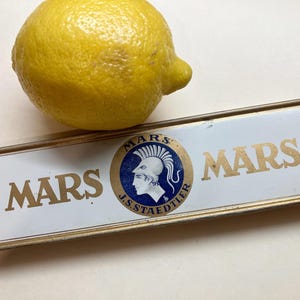 May include: A yellow lemon rests beside a rectangular, vintage Mars pencil case. The case is white with gold lettering and a circular emblem featuring a Roman soldier. The case is a product of J.S. Staedtler.