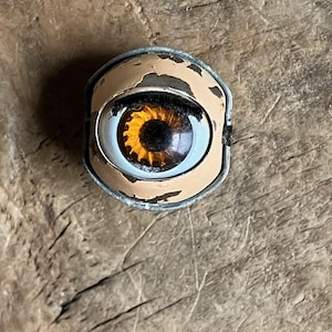 May include: A vintage, weathered eye-shaped object with a brown iris and a light blue sclera. The eye is set in a metal frame with peeling beige paint. The object rests on a rough, wooden surface.