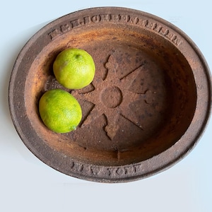 May include: A weathered, round, rusty metal bowl with the text "MELFORD IRON WORKS NEW YORK" around the rim. Two green limes rest inside the bowl, which has a sunburst design in the center.