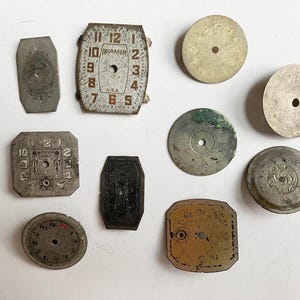 May include: A collection of nine vintage watch dials in various shapes and metallic finishes. The assortment includes rectangular, square, and circular faces, some featuring numerical markings and the text Ingraham U.S.A. on a rectangular dial.