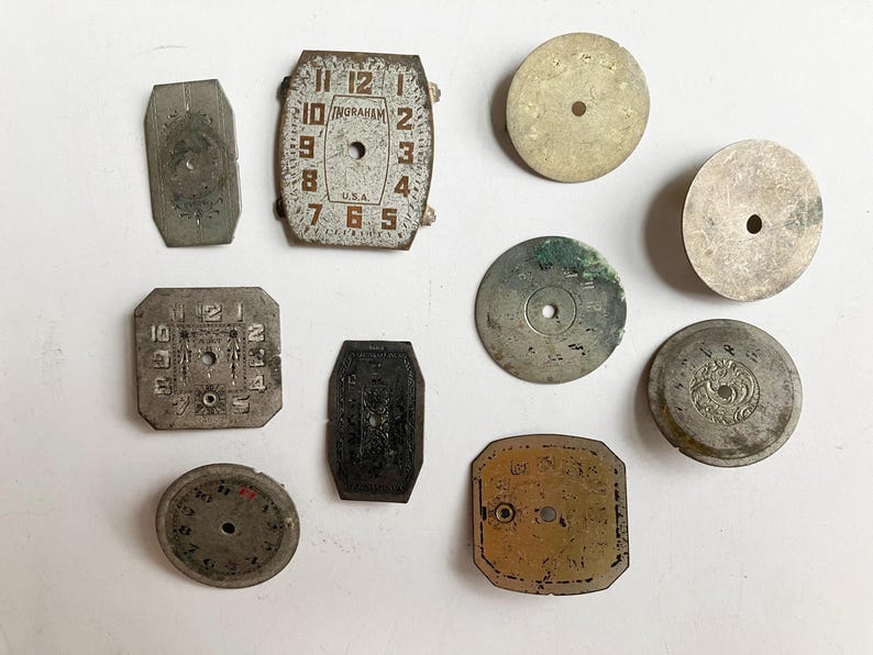 May include: A collection of nine vintage watch dials in various shapes and metallic finishes. The assortment includes rectangular, square, and circular faces, some featuring numerical markings and the text Ingraham U.S.A. on a rectangular dial.