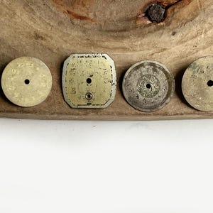 May include: Four vintage watch dials arranged in a row. Three are circular and one is square with rounded corners. All dials are metallic, showing signs of age and wear, and each features a central hole for hands. The surfaces are weathered.