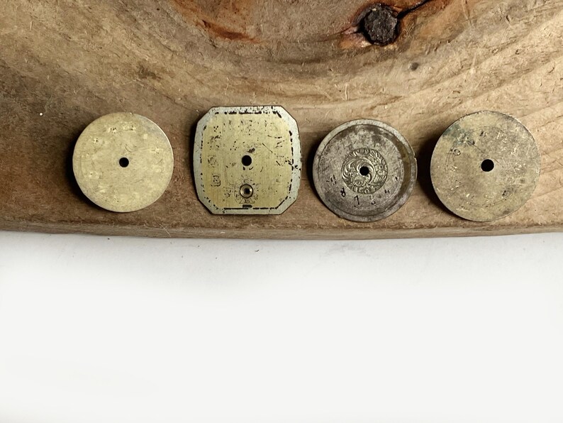 May include: Four vintage watch dials arranged in a row. Three are circular and one is square with rounded corners. All dials are metallic, showing signs of age and wear, and each features a central hole for hands. The surfaces are weathered.