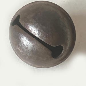 May include: A spherical metal bell with a dark, weathered, and oxidized brown finish. The bell features a prominent, elongated vertical slit across its center, which widens into circular openings at both ends, creating a classic jingle bell design.