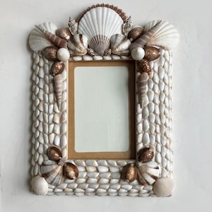 May include: A rectangular photo frame decorated with various seashells and small white pebbles. The frame features a central opening for a photo, with a brown border. The frame is adorned with white, brown, and gold seashells.